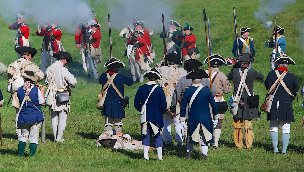 Revolutionary War reenactors engaged in battle on a green field.